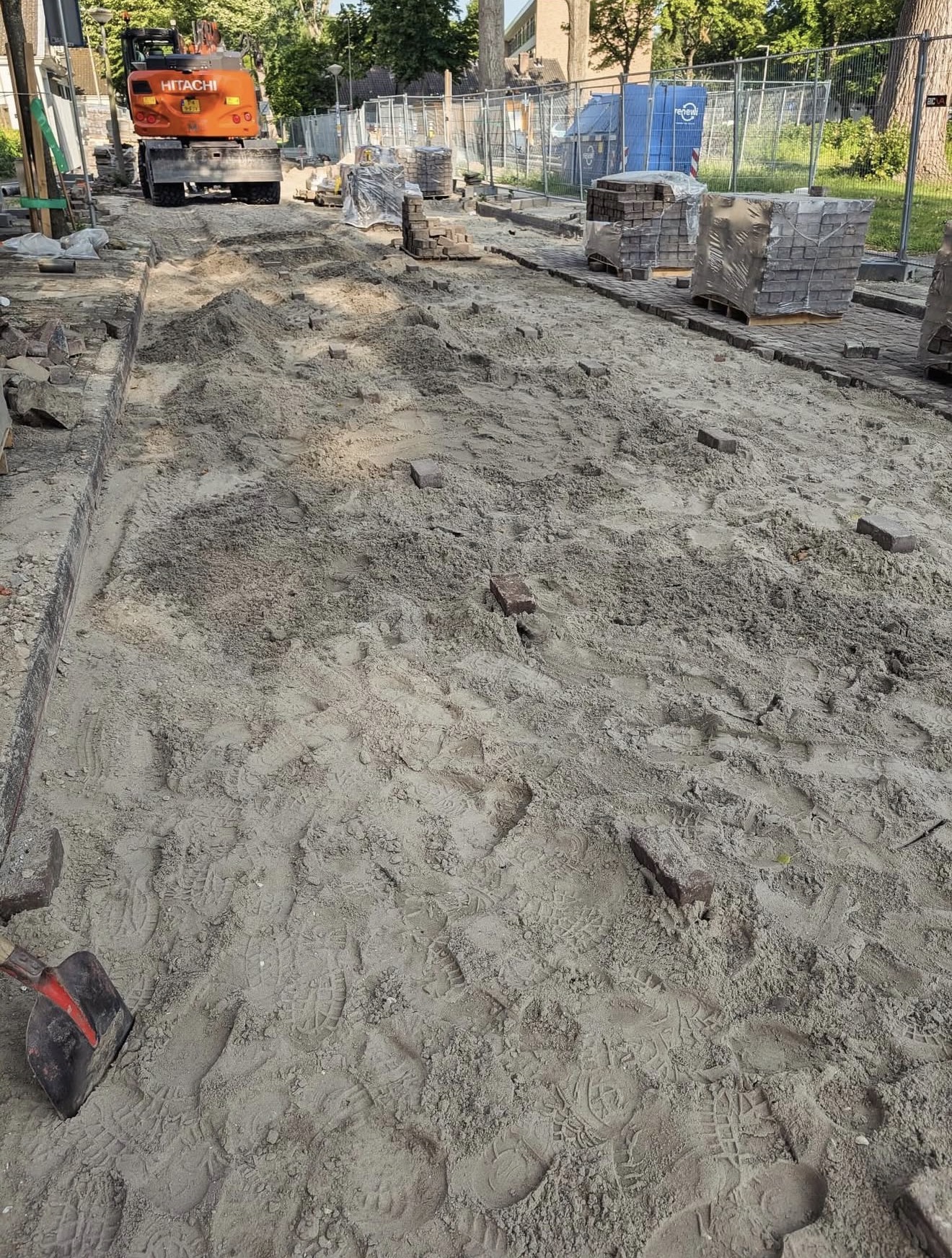 Opengebroken straat met Hitachi graafmachine en pallets klinkers
